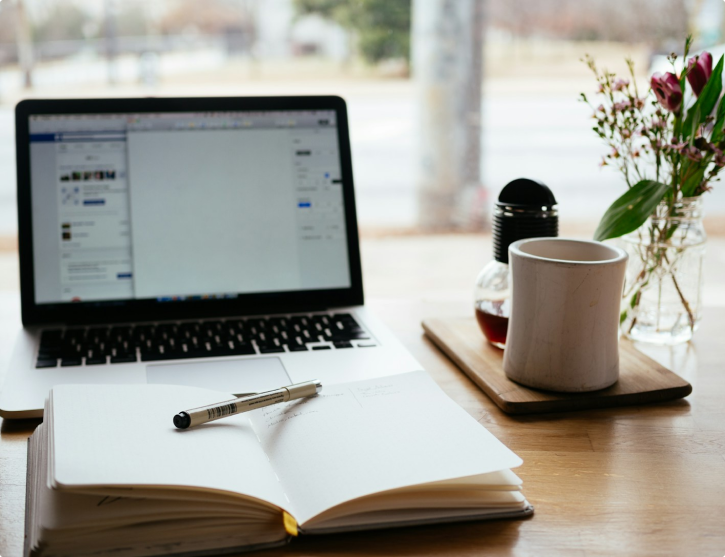 A bright, organized workspace featuring an open laptop, a notebook with a pen, a coffee mug, and a small vase of flowers on a wooden desk.