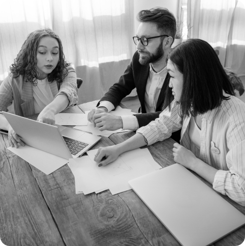 Three colleagues in business attire sit around a wooden table, focused on a laptop screen and scattered paperwork.